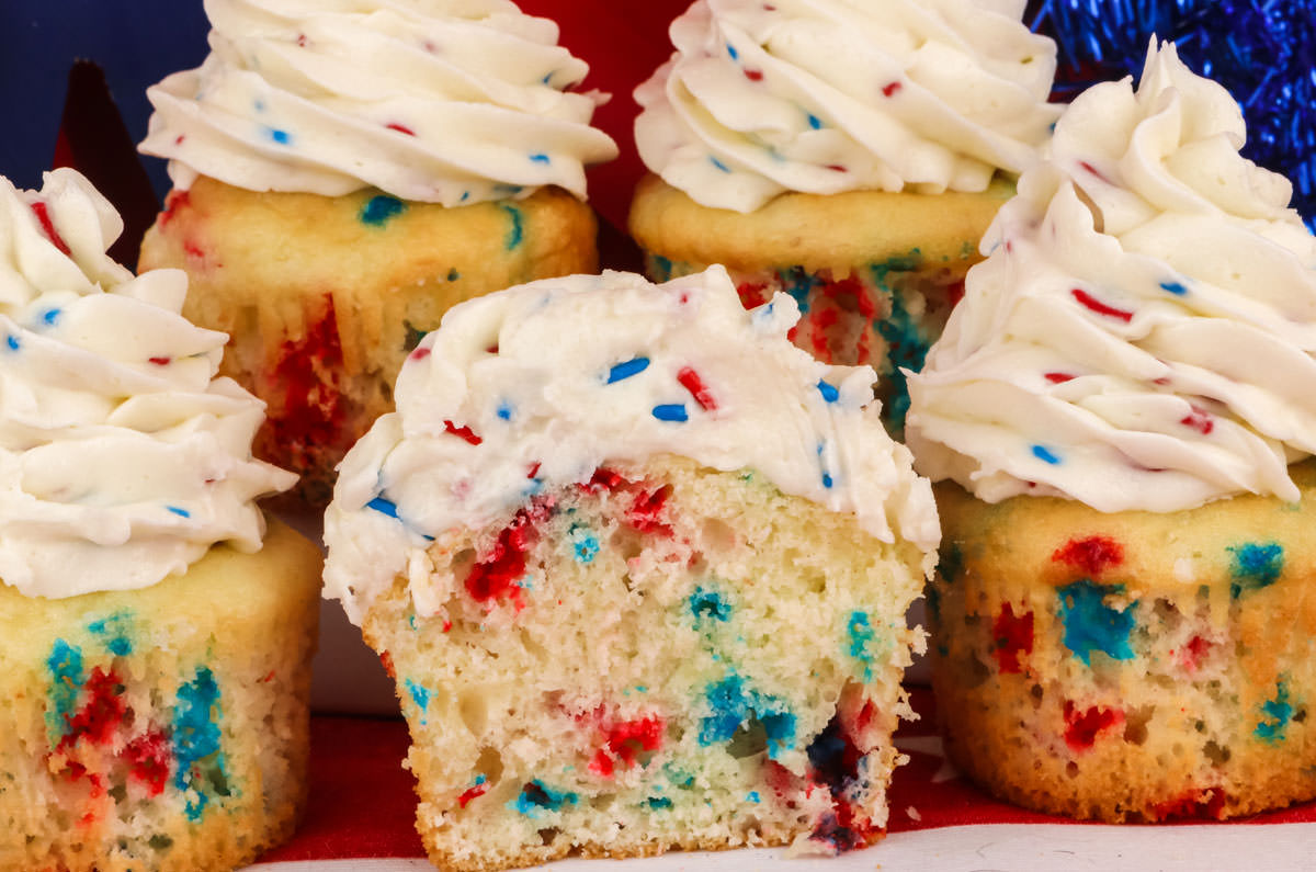 Close up image of five Red White and Blue Confetti Cupcakes sitting on a white table with one of the cupcakes cut in half so you can see the inside of the cupcake.