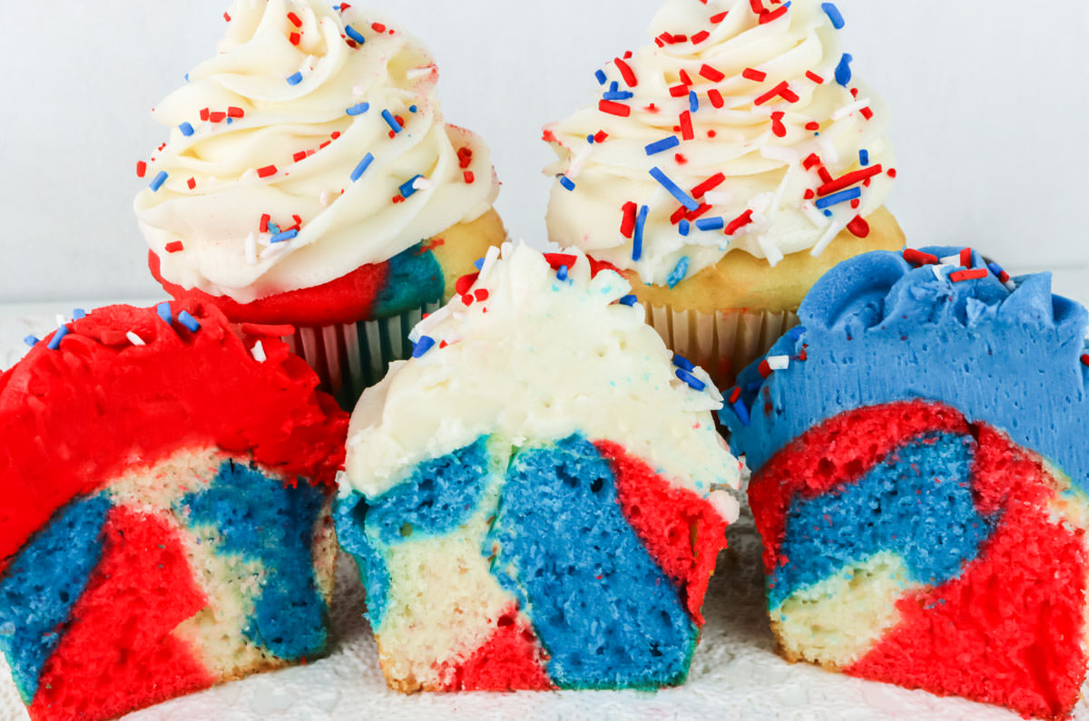 Close up on five Patriotic Marble Cupcakes sitting on a white table, with three of the cupcakes cut in half to show the marbling effect inside the cupcake.