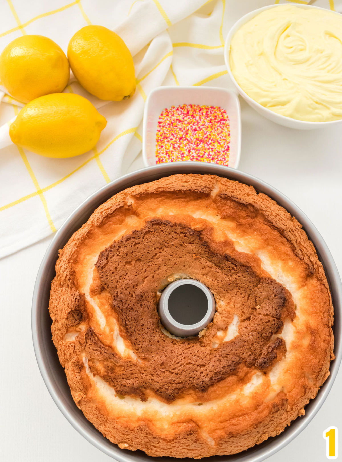 Closeup of an Angel Food Cake just out of the oven and still in the cake pan sitting on a white surface in front of a ramekin of sprinkles and a bowl of lemon frosting.