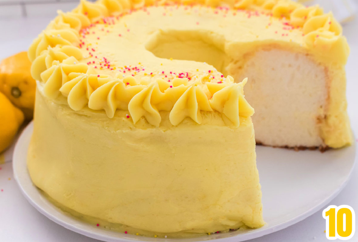 Closeup on an Angel Food Cake with Lemon Frosting sitting on a white table with a piece taken out of it and lemons off to the side.