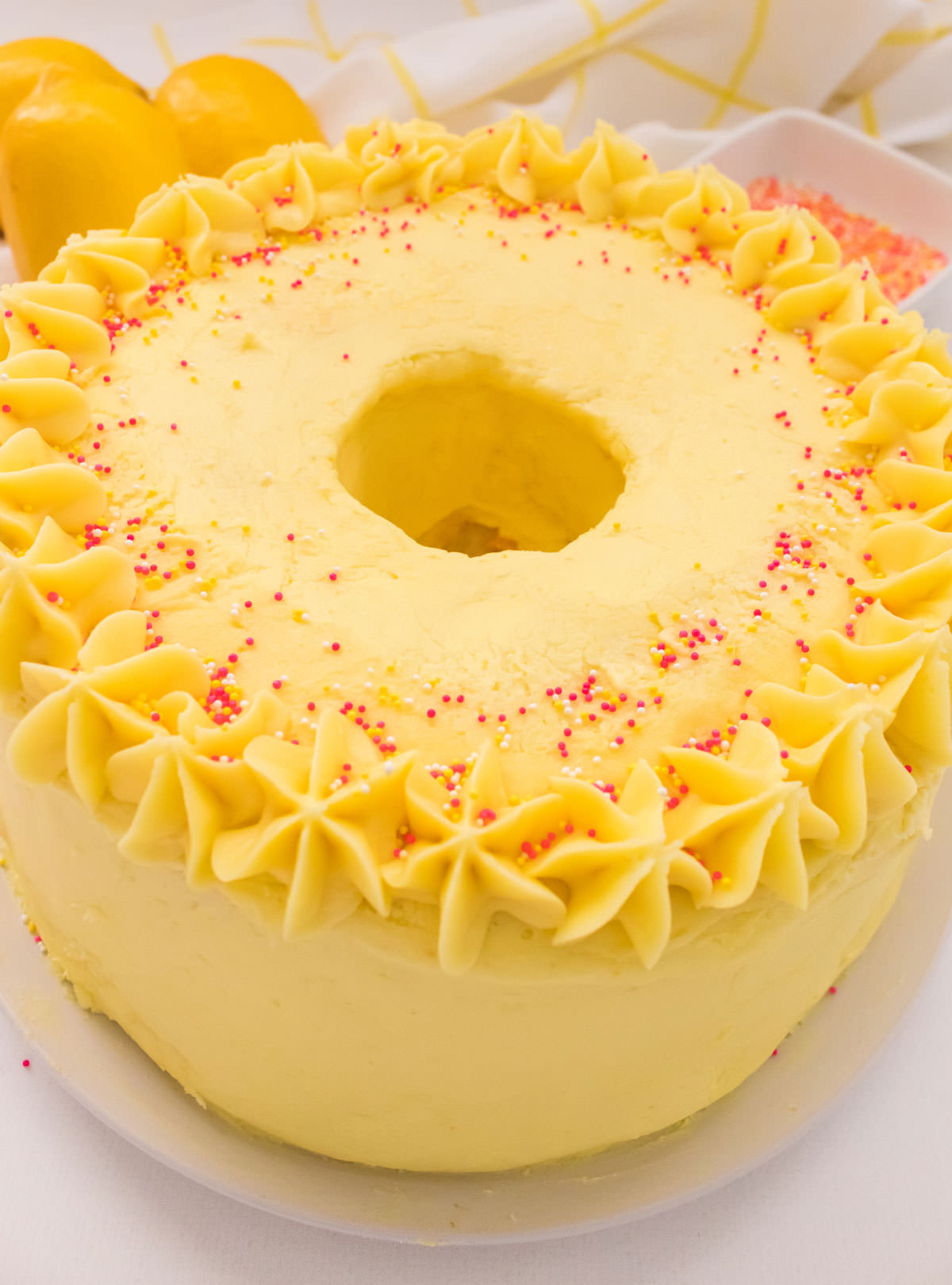 Closeup on an Angel Food Cake with Lemon Frosting sitting on a white cake plate on a white table with lemons in the background.