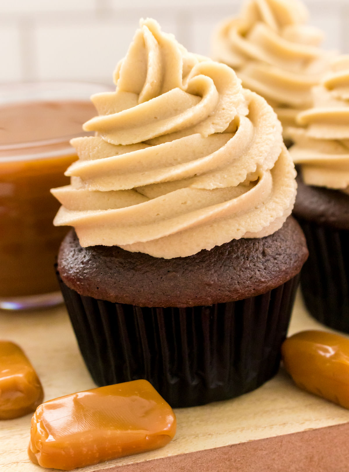 Closeup on two chocolate cupcakes frosted with The Best Caramel Buttercream Frosting sitting next to a bowl of caramel sauce.