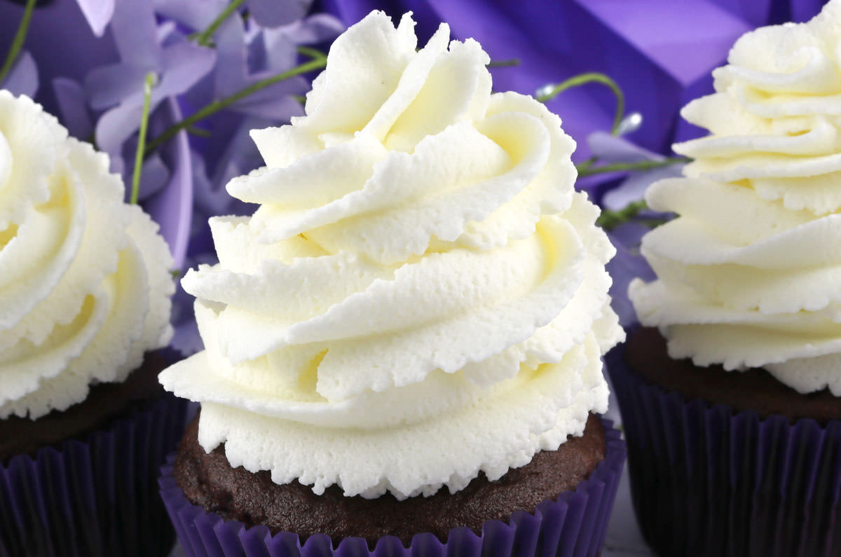 Closeup on three cupcakes topped with The Best Whipped Cream Frosting sitting on a white surface in front of a purple background.