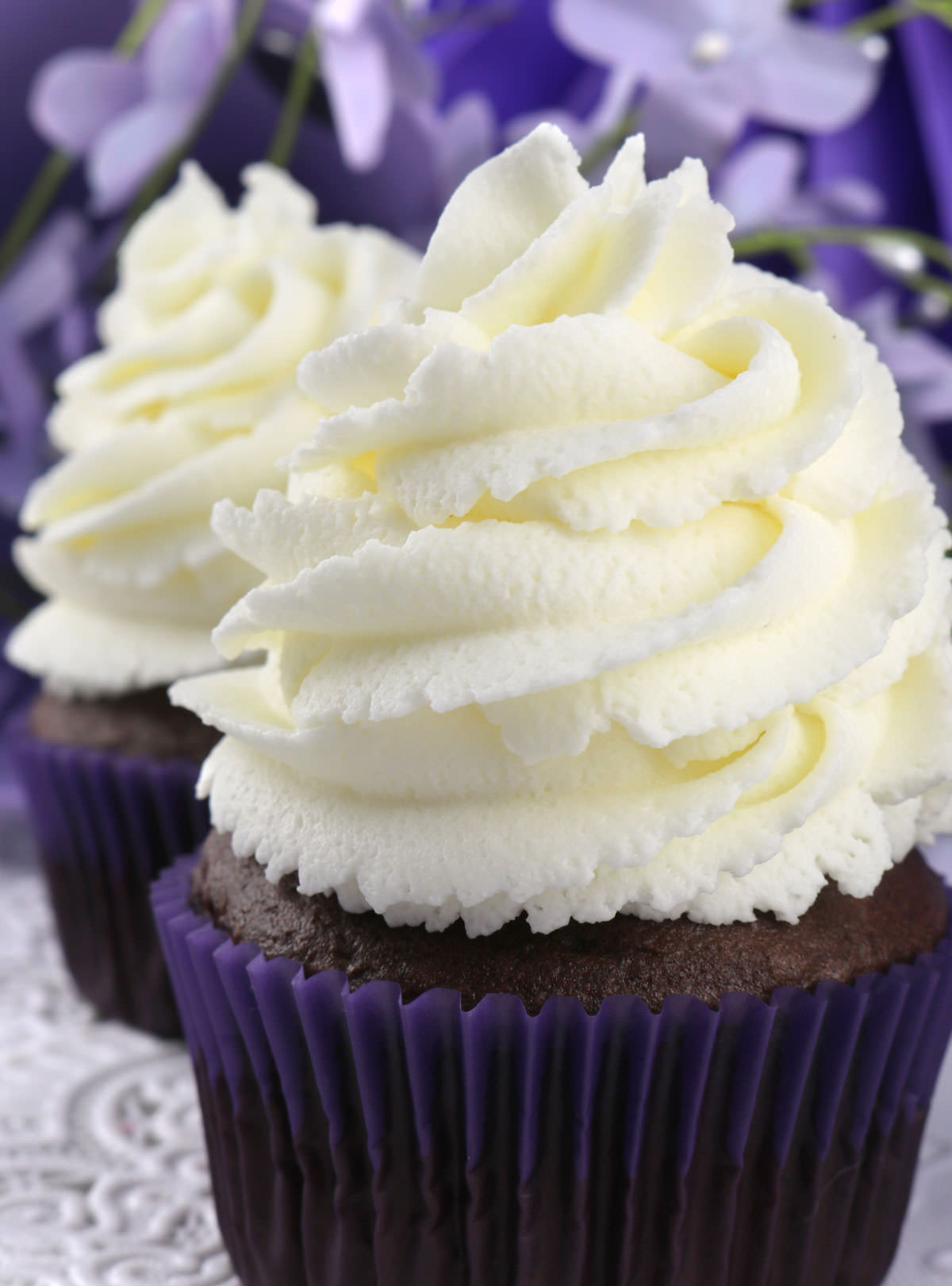 Closeup on two cupcakes topped with The Best Whipped Cream Frosting sitting on a white table in front of purple flowers.