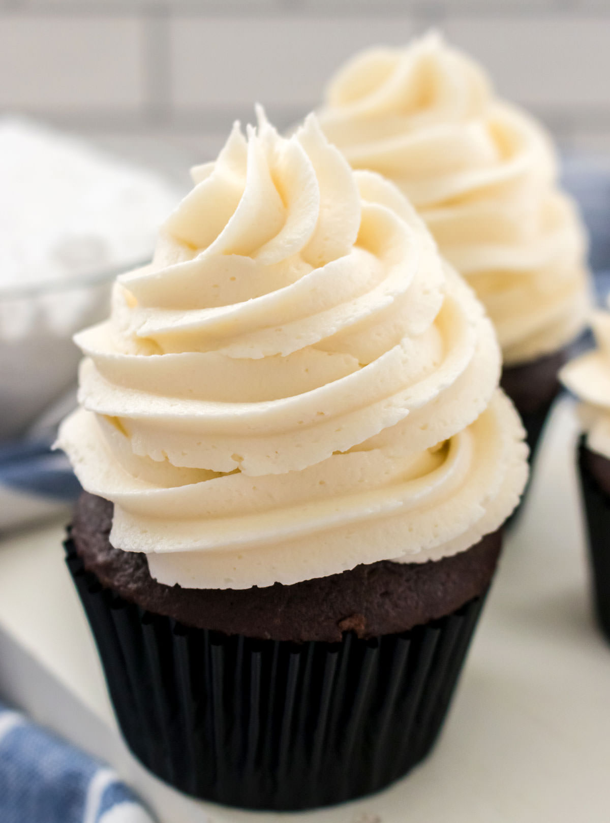 Closeup on two cupcakes topped with The Best Whipped Vanilla Frosting sitting next to a bowl of powdered sugar.