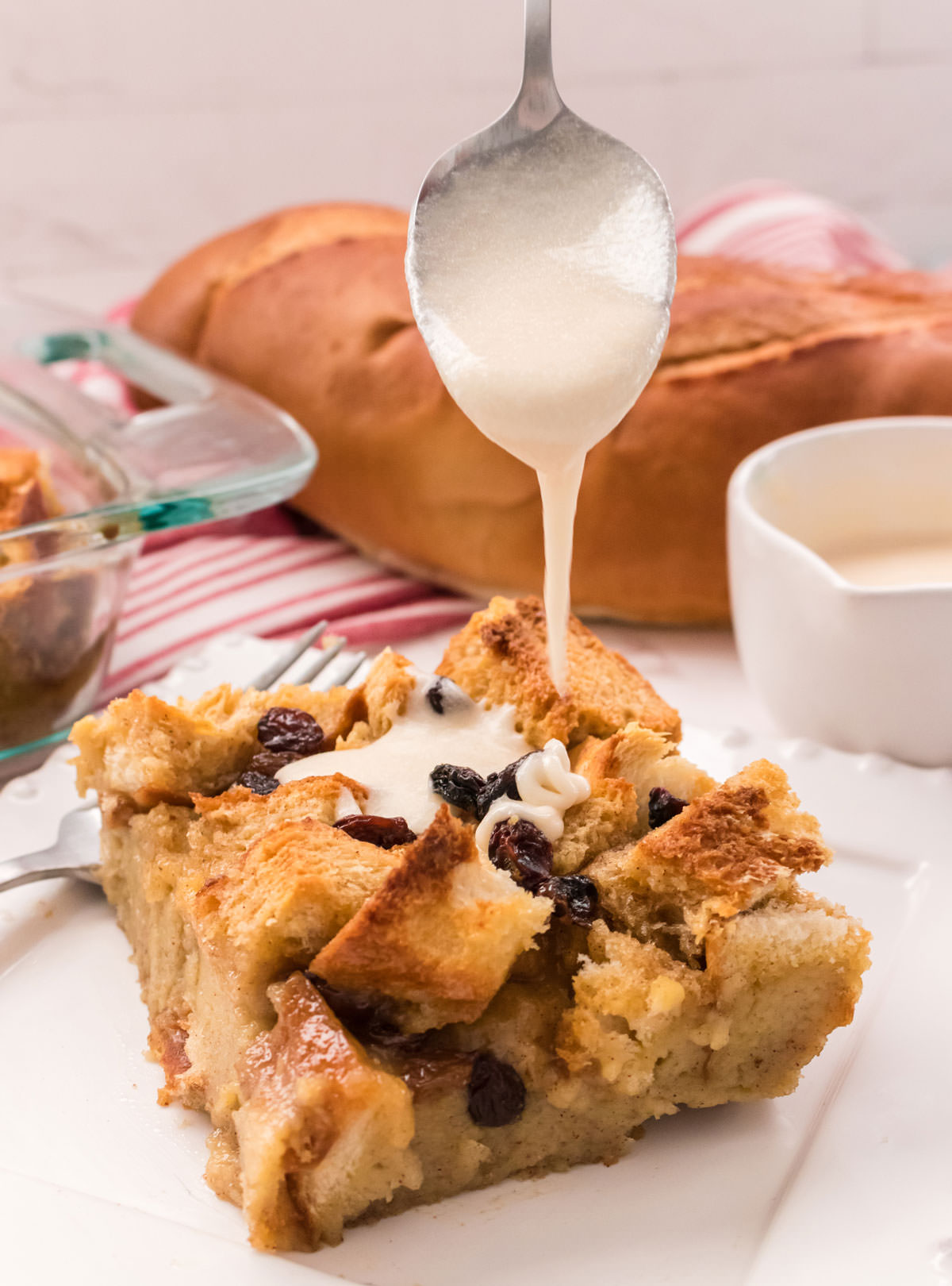 Closeup of a piece of Bread Pudding sitting on a white plate with a spoon dropping vanilla sauce onto the top of the dessert.