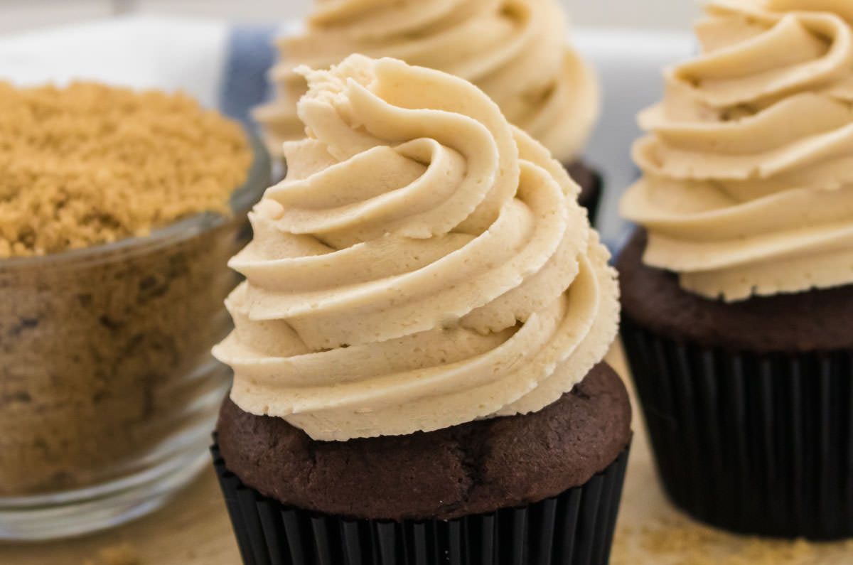 Closeup on three cupcakes topped with The Best Brown Sugar Buttercream Frosting next to a glass bowl filled with Brown Sugar.