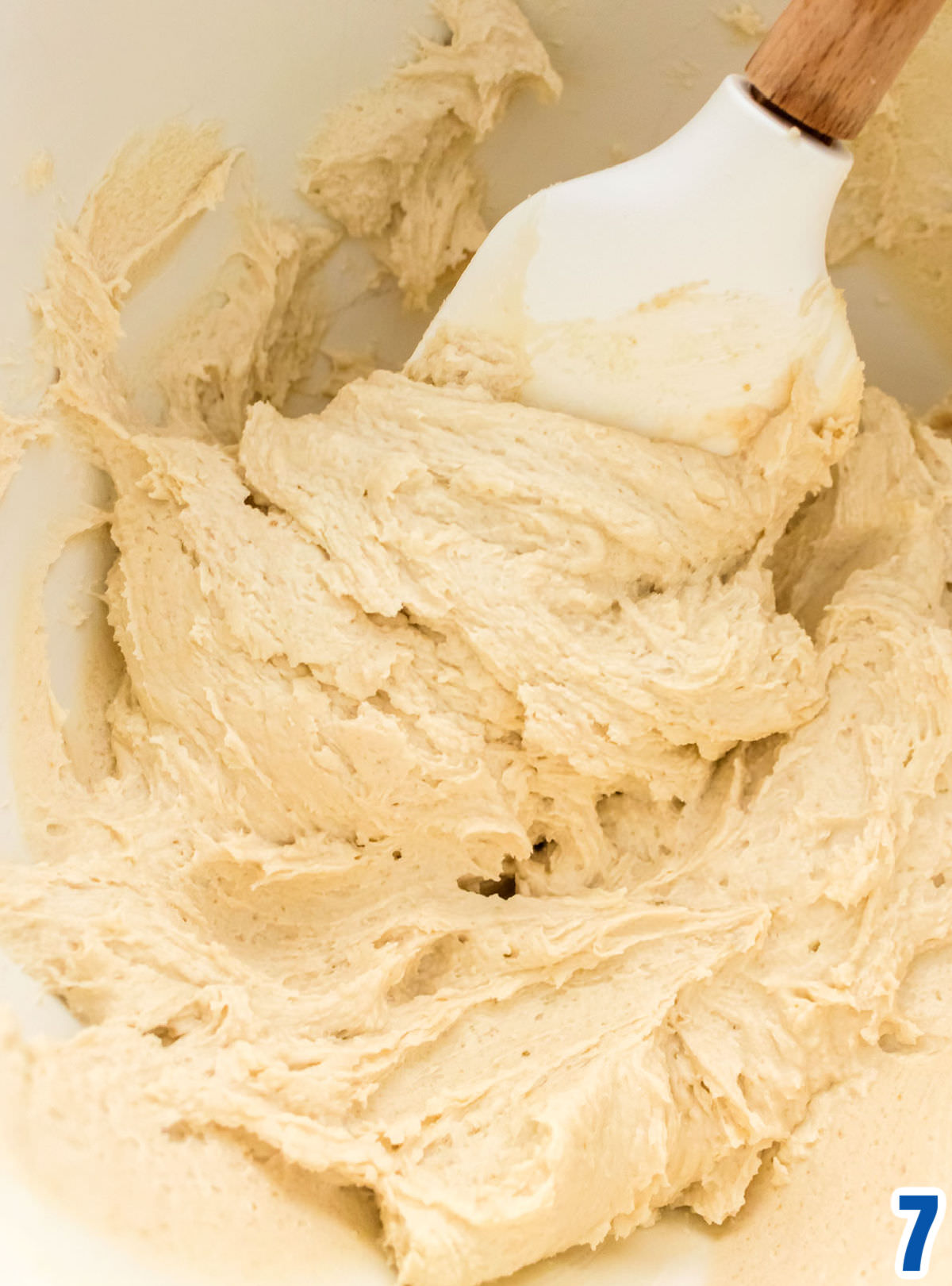 Closeup on a white mixing bowl filled with Brown Sugar Icing  and a white wooden spatula.