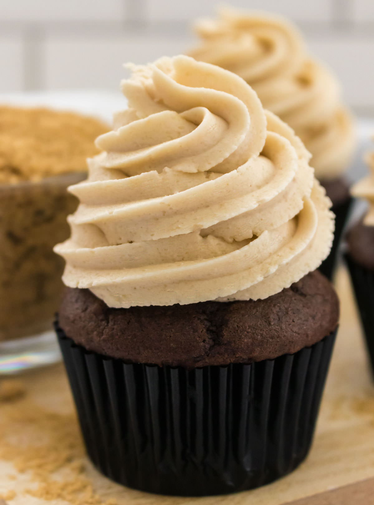 Closeup on a chocolate cupcake topped with The Best Brown Sugar Buttercream Frosting sitting on a cutting board next to a glass bowl filled with Brown Sugar.