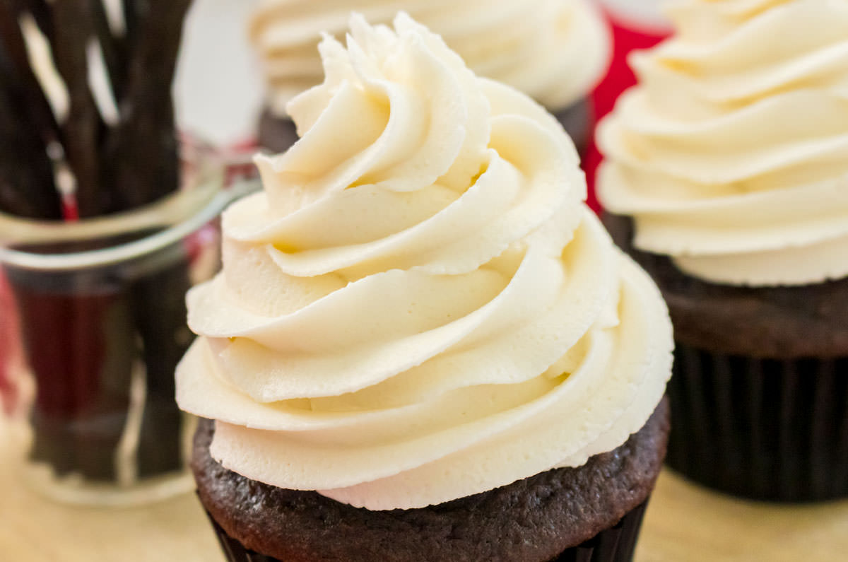 Closeup on a cupcake topped with The Best Buttercream Frosting sitting in front of other cupcakes and a jar filled with Vanilla Beans.