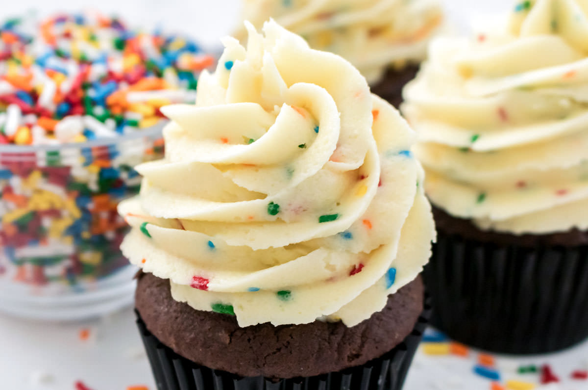 Closeup on three cupcakes frosted with The Best Cake Batter Buttercream Frosting sitting next to a glass bowl filled with sprinkles.
