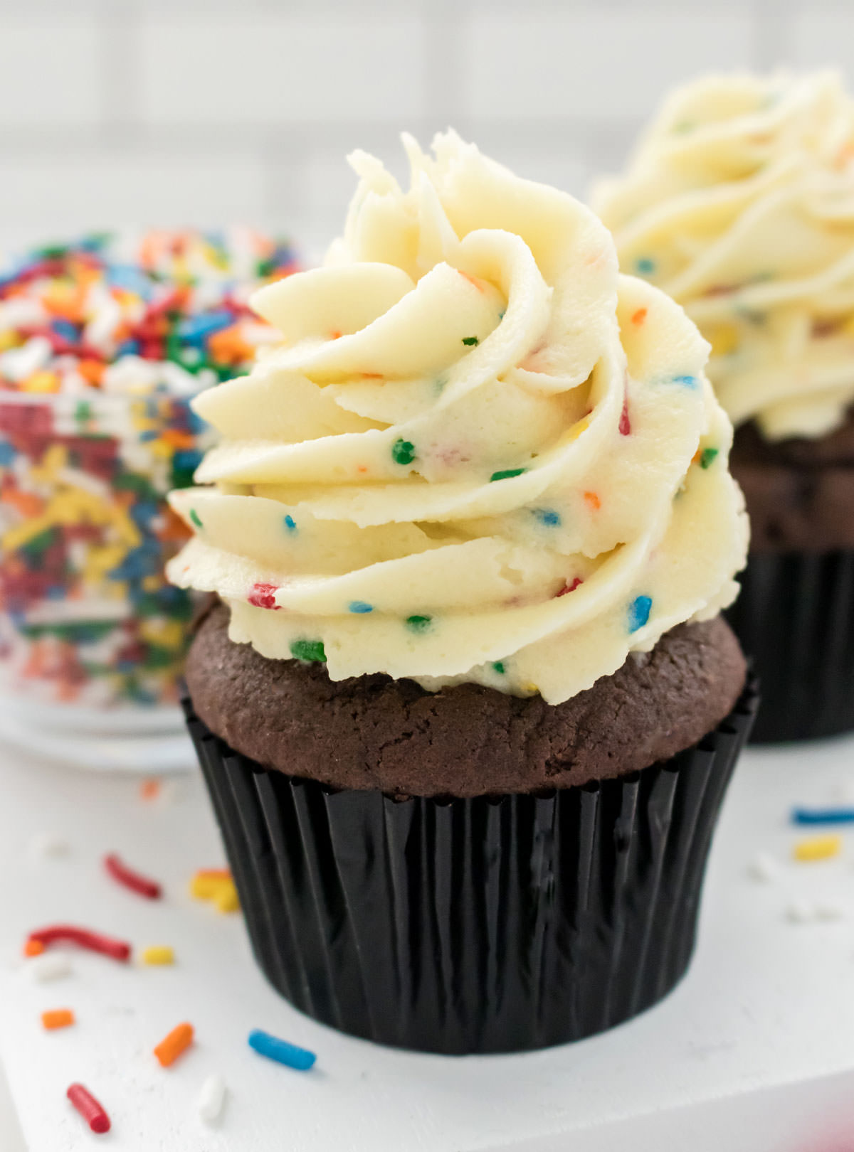 Closeup on a chocolate cupcake topped with The Best Cake Batter Buttercream Frosting sitting next to a glass bowl filled with sprinkles.