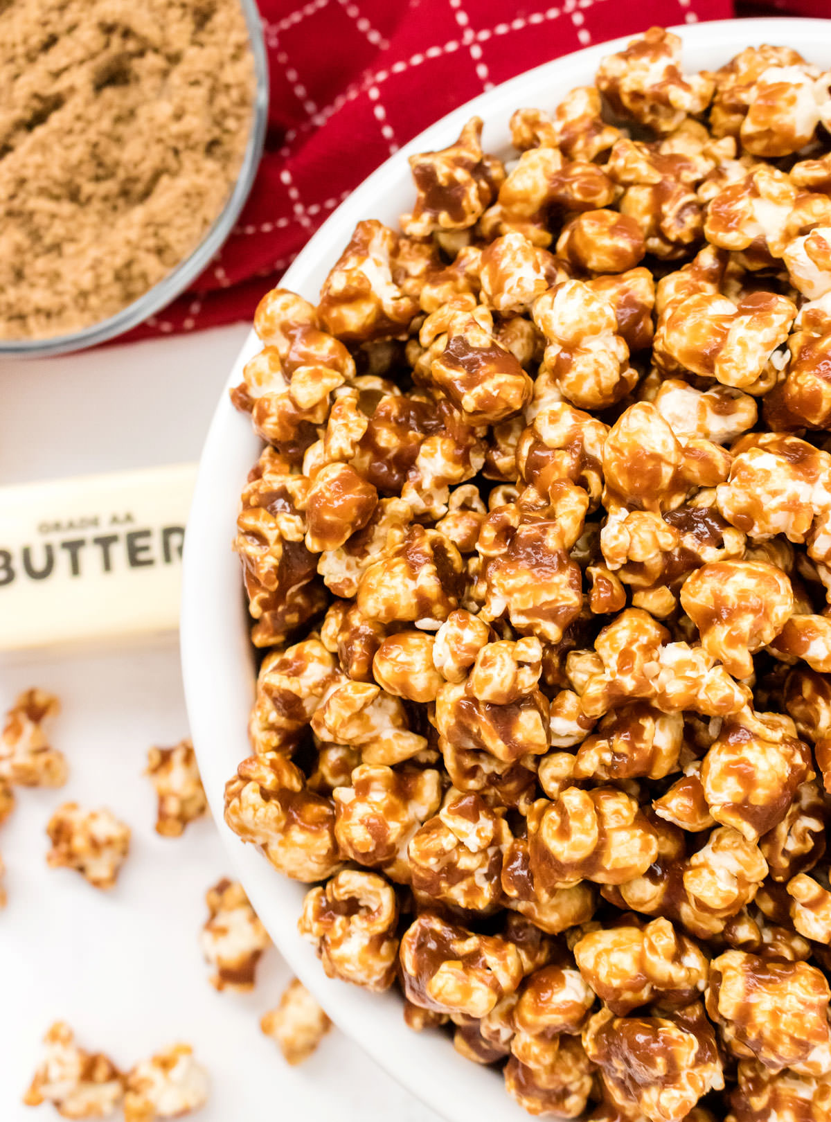 Overhead closeup on a white serving bowl filled with Easy Homemade Caramel Corn sitting next to a bowl of brown sugar, a stick of butter and a red towel.