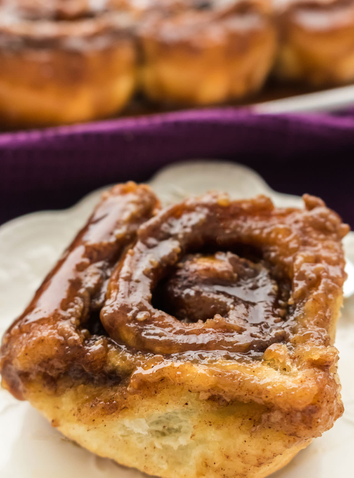 A Caramel Sticky Bun sitting on a white plate in front of a platter full of the rest of the pastries.