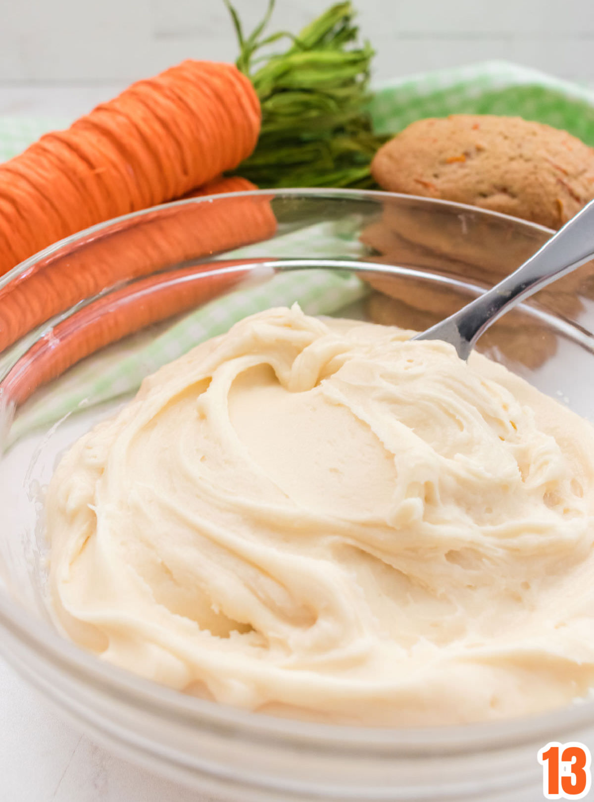 Closeup of a clear mixing bowl filled with Cream Cheese Frosting sitting in front of a stack of cookies and carrot decorations.