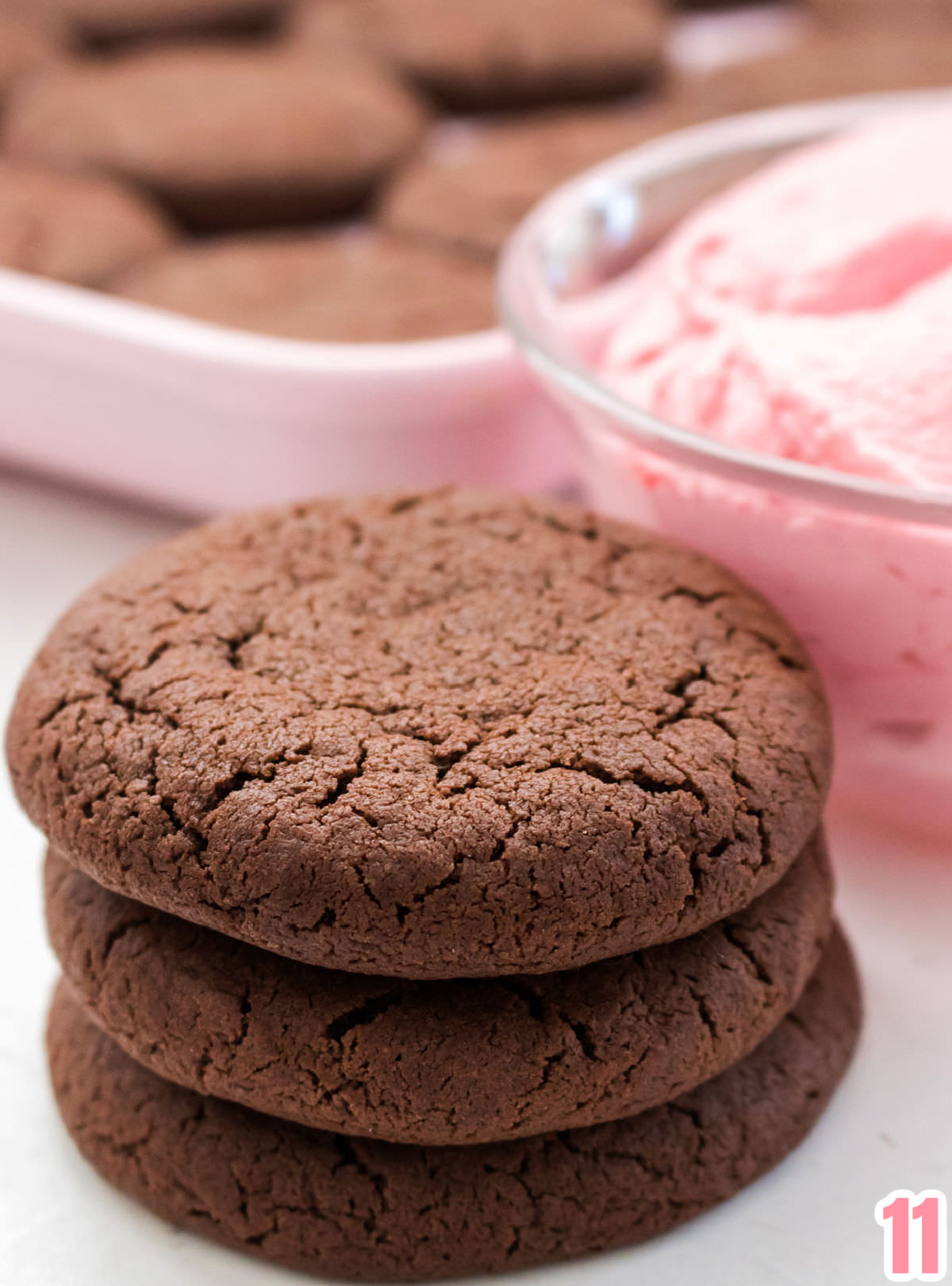 Close up of a stack of three chocolate cookies sitting next to a bowl of Raspberry Frosting and in front of a cookie sheet filled with chocolate cookies.
