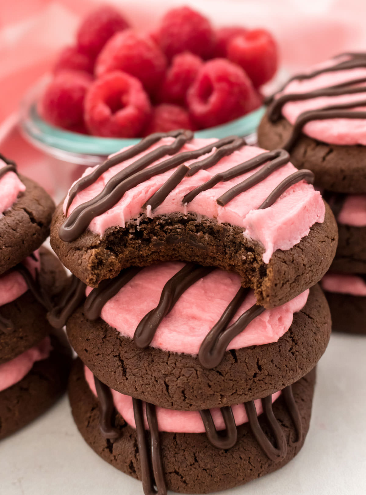 Closeup on a stack of Chocolate Cookies with Raspberry Frosting sitting on a table in front of a glass bowl filled with fresh Raspberries.