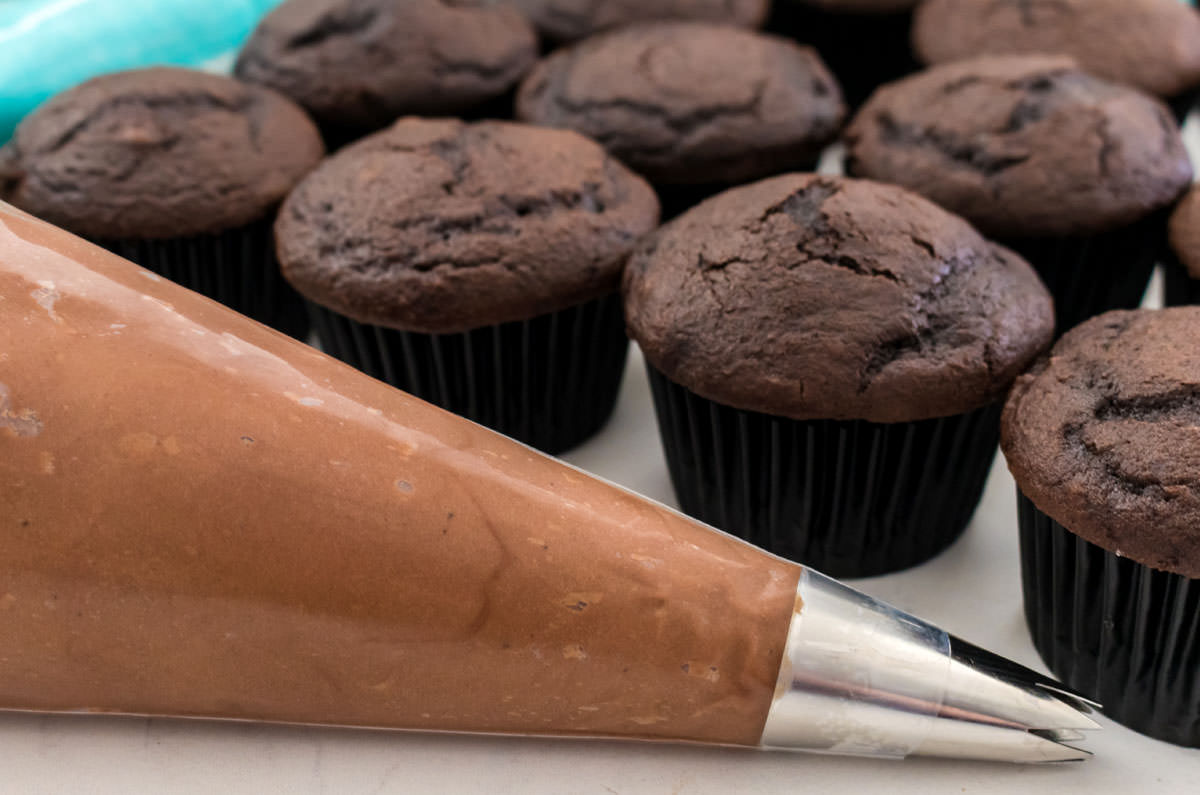 A decorating bag filled with Homemade Chocolate Cream Cheese Icing sitting on a white table in front of a batch of unfrosted chocolate cupcakes.