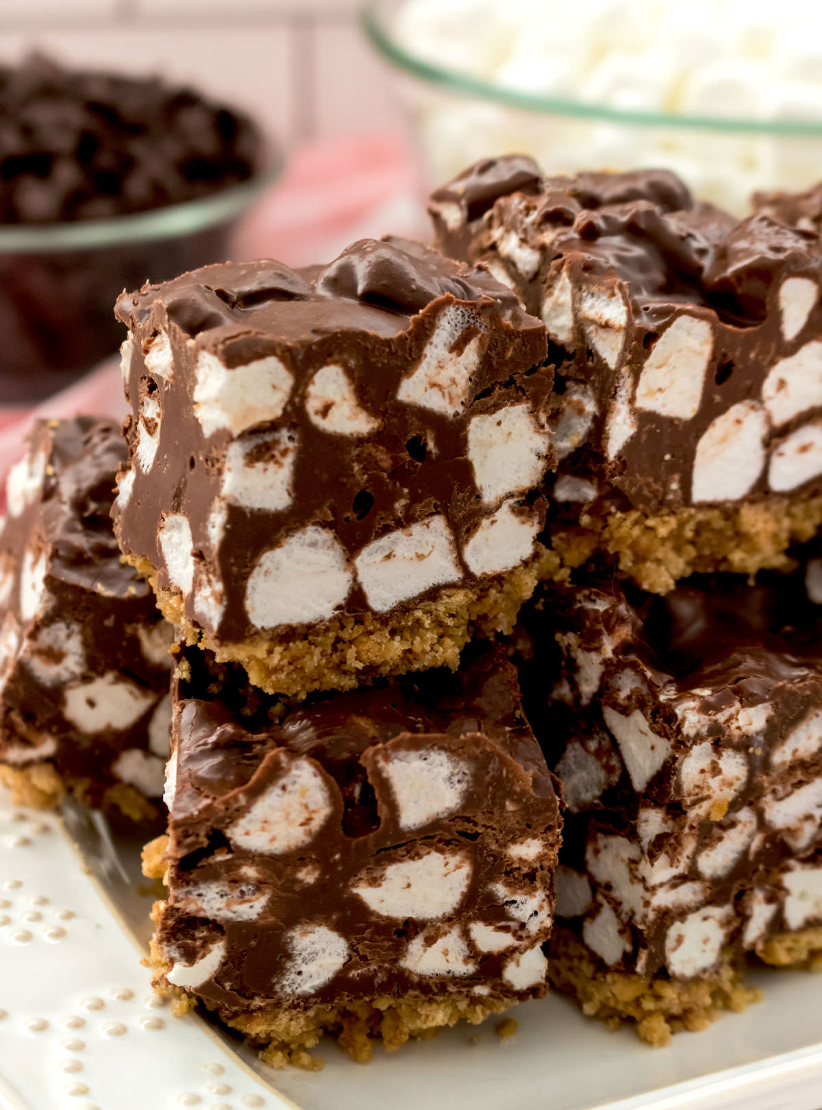 Closeup on a stack of Chocolate Marshmallow Bars sitting on a white serving platter in front of glass bowls filled with chocolate chips and mini marshmallows.