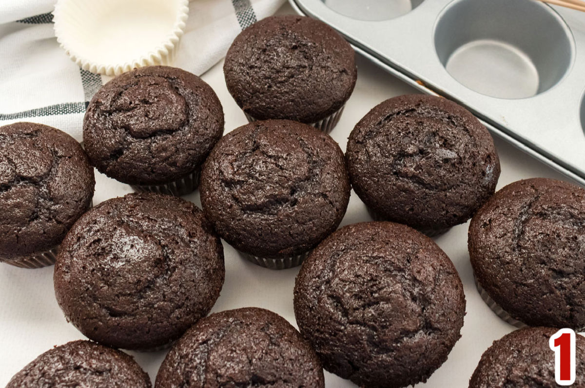 A dozen chocolate cupcakes sitting on a white table next to a cupcake pan.