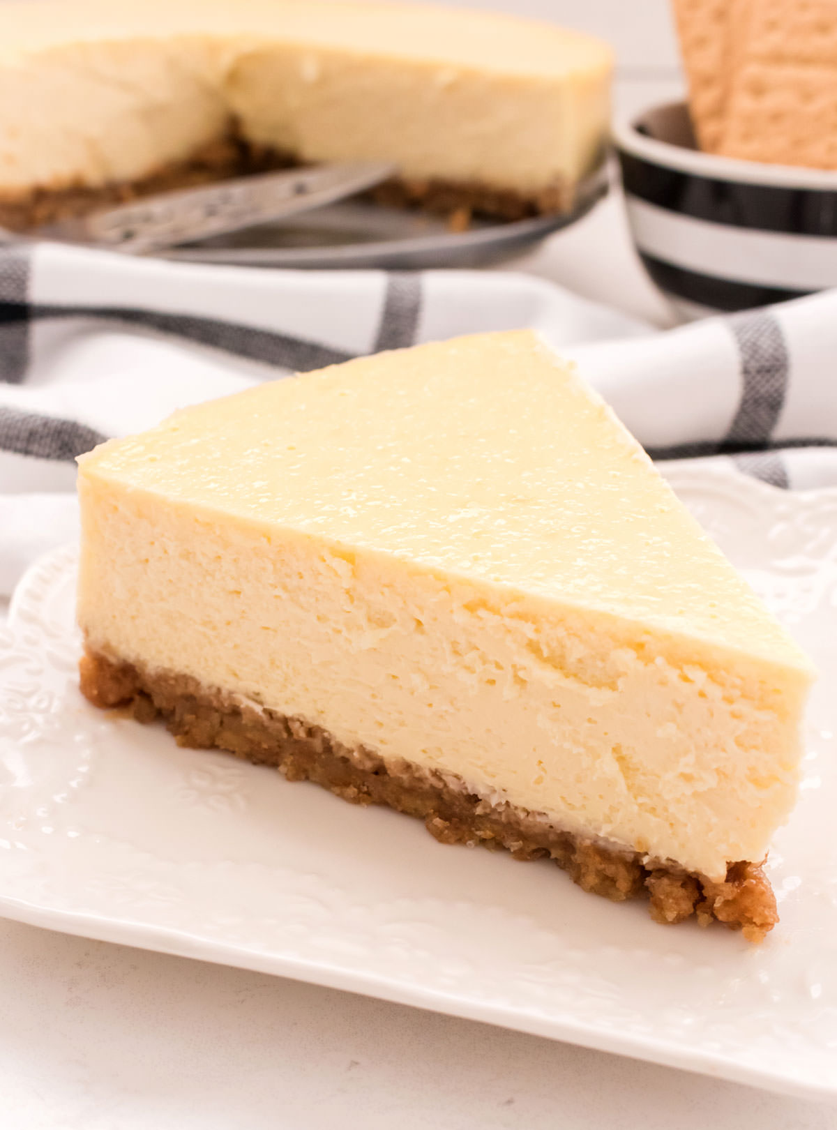 Closeup of a single piece of Classic Cheesecake sitting on a white plate in front of the remaining cheesecake in the background.