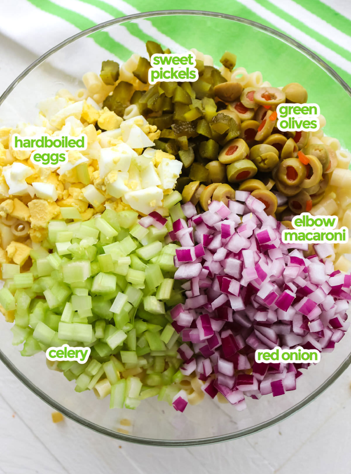 All the ingredients you will need to make Classic Macaroni Salad including short macaroni pasta, sweet pickels, green olives, red onion, celery, and hardboiled eggs.