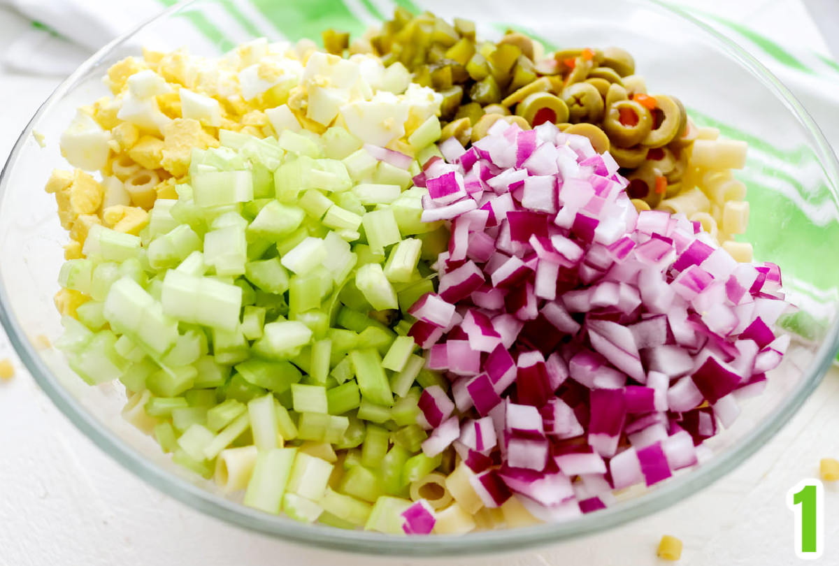 Clear glass bowl filled with cooked short macaroni, chopped olives, chopped red onion, chopped celery and chopped hard boiled eggs.