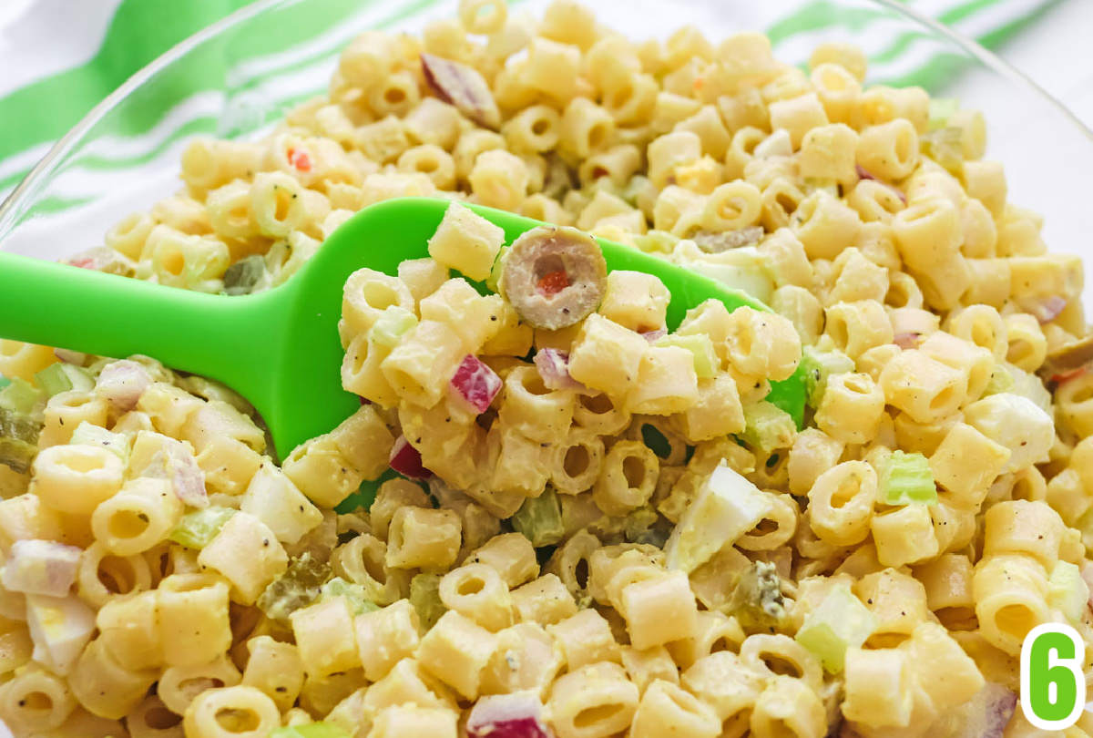 Closeup on a clear glass bowl filled with Macaroni Salad with a green spoon scooping out some of the salad.