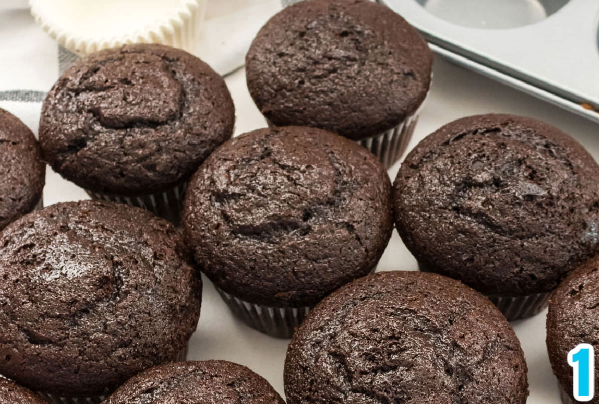 Closeup on a batch of chocolate cupcakes sitting on a white surface with a cupcake tin and cupcake liners in the background.