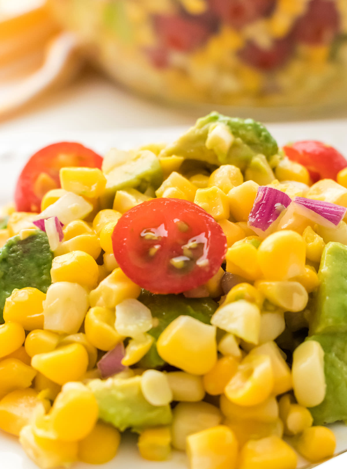 Closeup on a large helping of The Best Ever Summer Corn Salad sitting on a white plate on a white table in front of a glass bowl filled with Corn Salad.