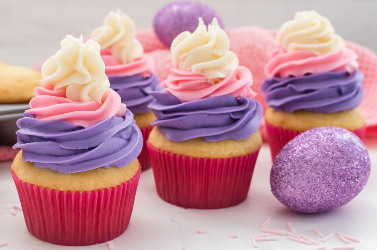 Closeup of four Easter Cupcakes sitting on a white surface next to an easter egg, a pink towel and a pan of vanilla cupcakes.