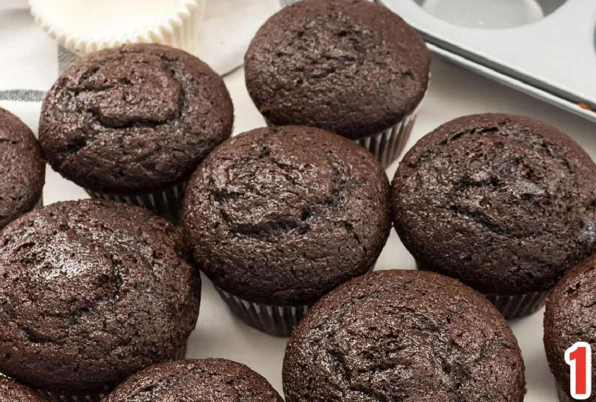 A dozen chocolate cupcakes sitting on a white surface in front of a cupcake and cupcake liners.