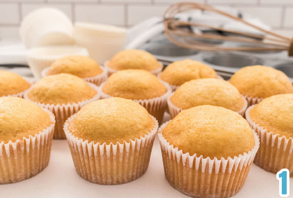 12 vanilla cupcakes sitting on a white surface in front of cupcake tins and a whisk.