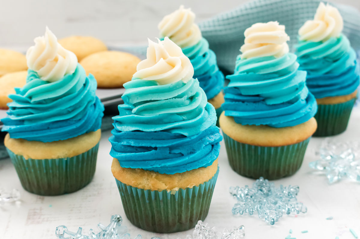 Closeup on five Frozen Ombre Swirl Cupcakes sitting on a white table surrounded by snowflake decorations, cupcakes and a blue table linen.