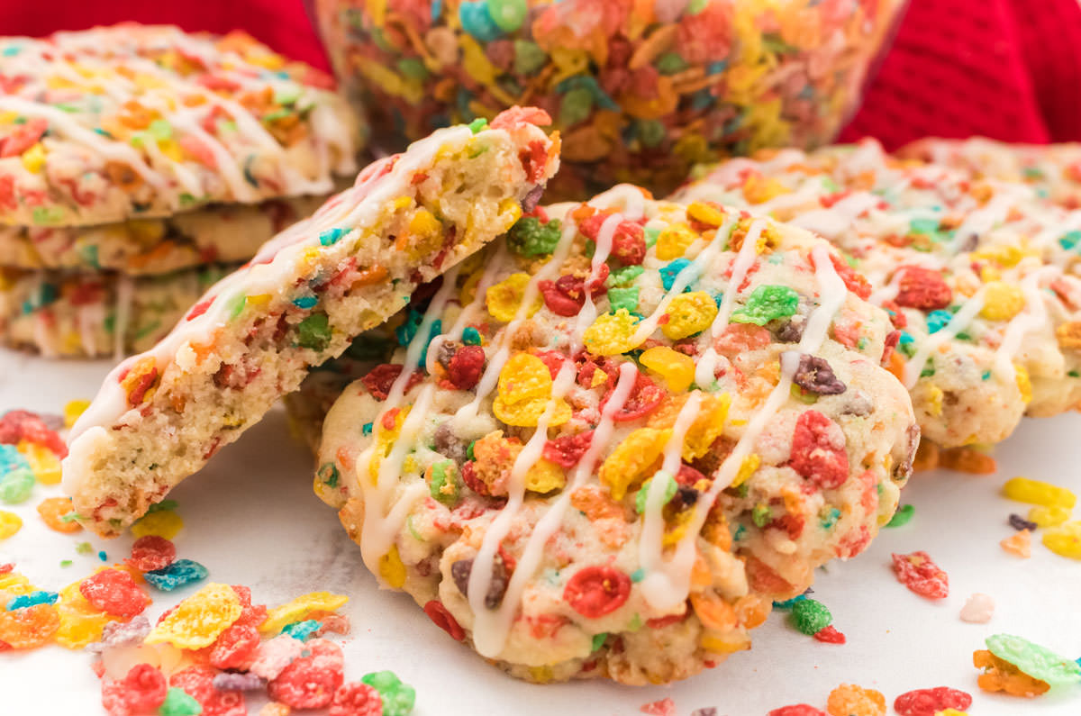Closeup on a batch of Fruity Pebble Sugar Cookies laying on a white table surrounded by Fruity Pebble Cereal.