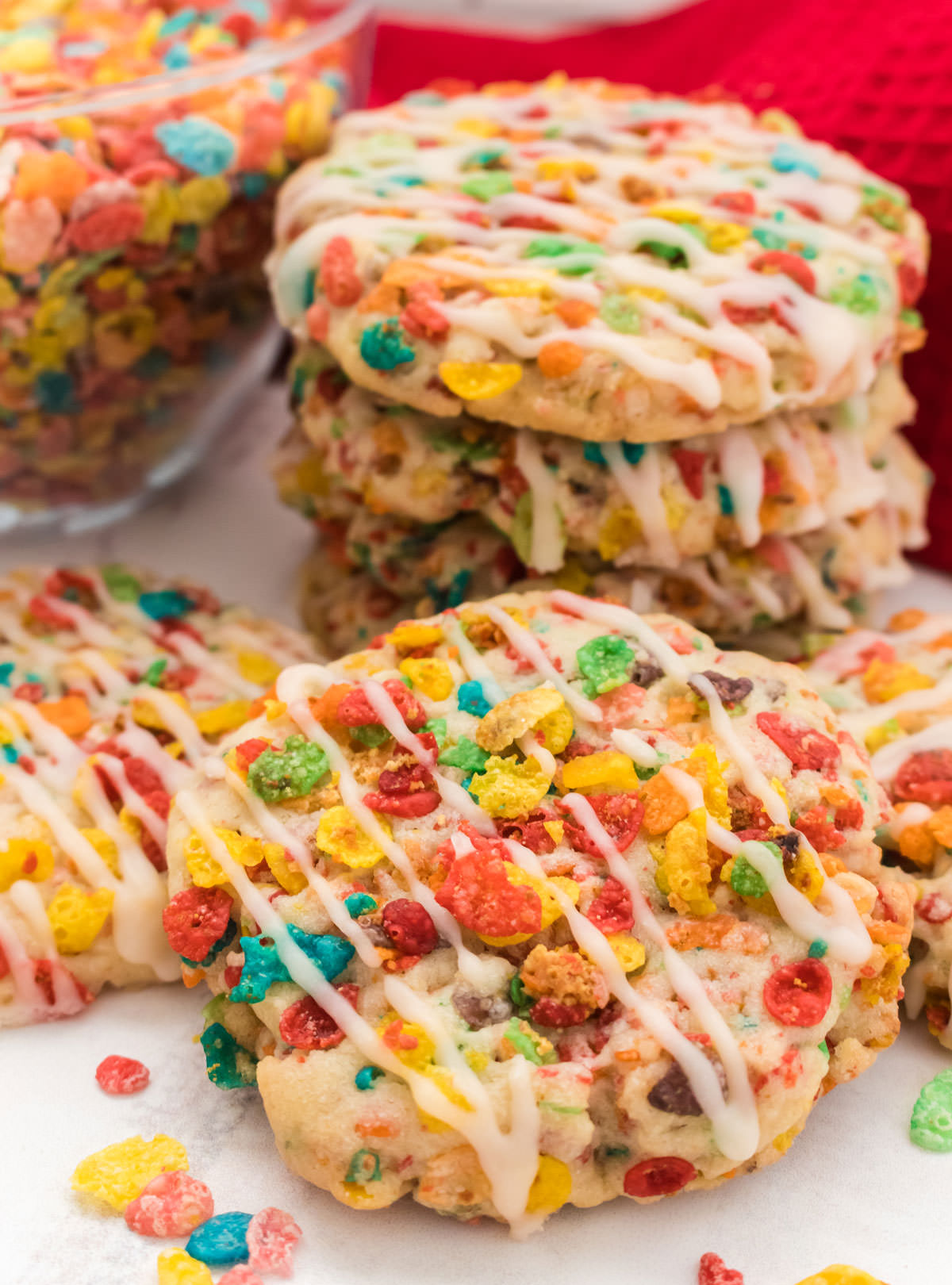 Closeup on a Fruity Pebbles Sugar Cookie propped up against another stack of cookies and in front of a glass bowl filled with. Fruity Pebbles Cereal.