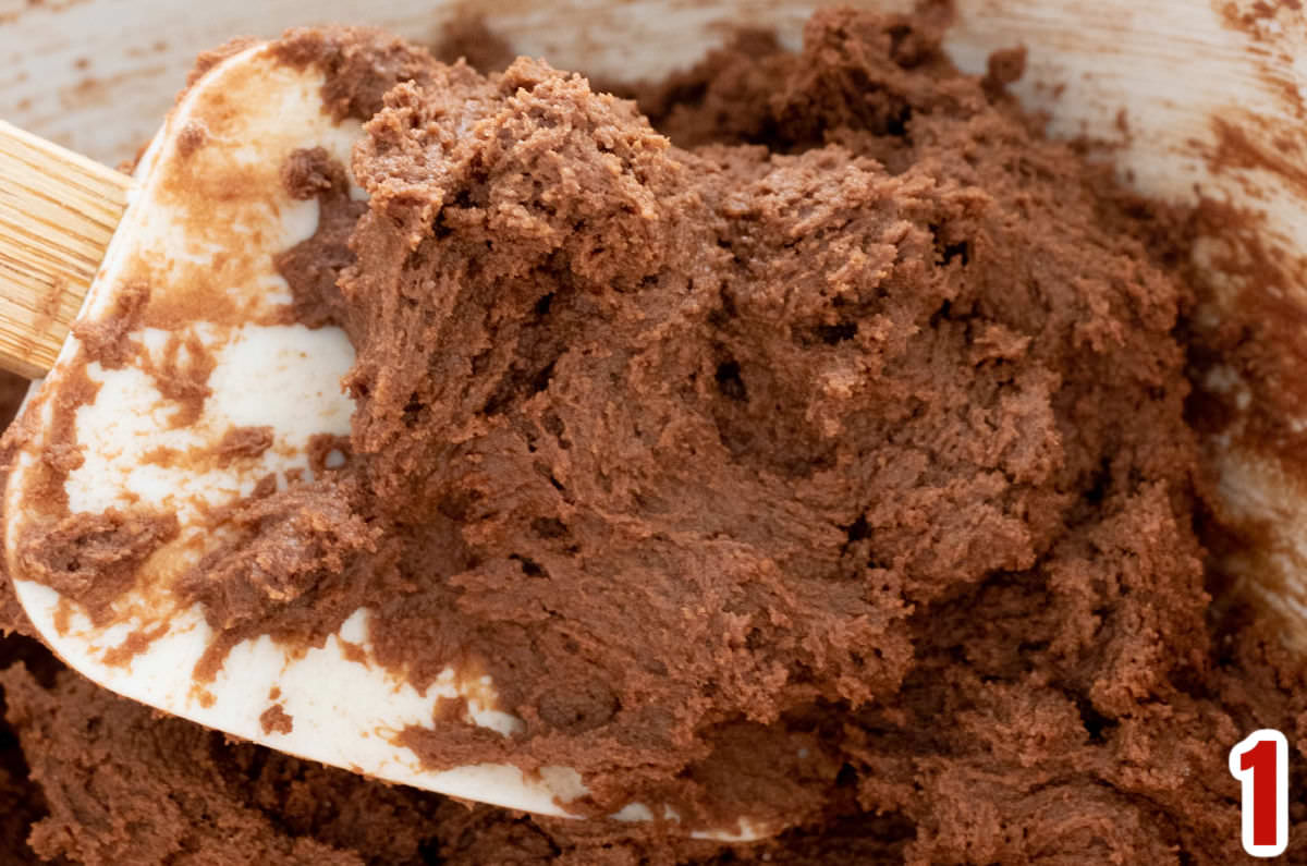 Closeup on a white bowl filled with chocolate cookie dough and a white wooden spatula.