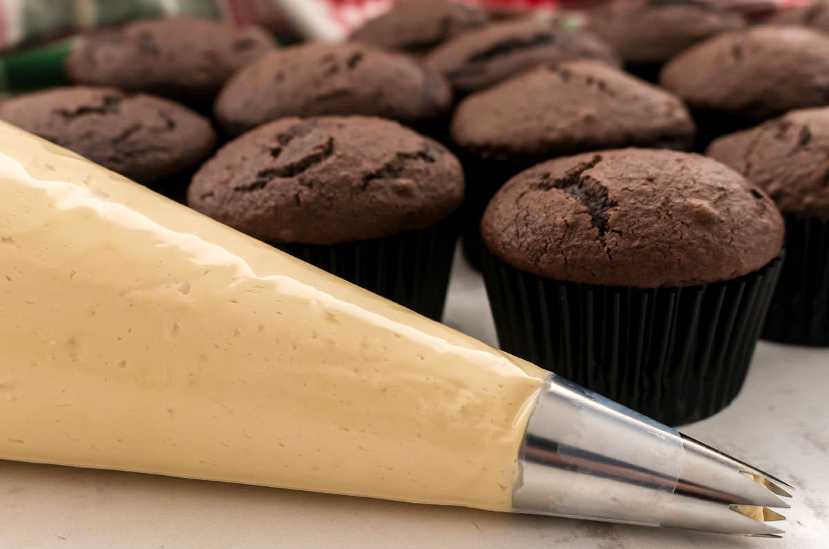 Closeup on a decorating bag filled with Gingerbread Frosting sitting in front of a batch of unfrosted chocolate cupcakes.