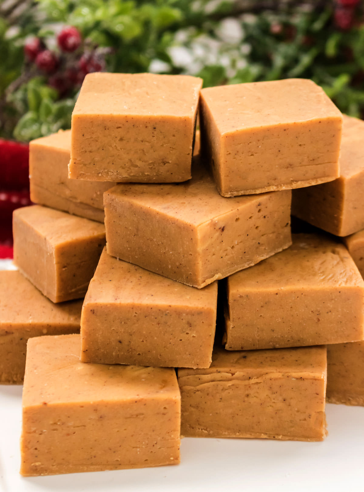 Closeup on a stack of Gingerbread Fudge pieces sitting on a white table in front of holiday greenery and holly leaves.