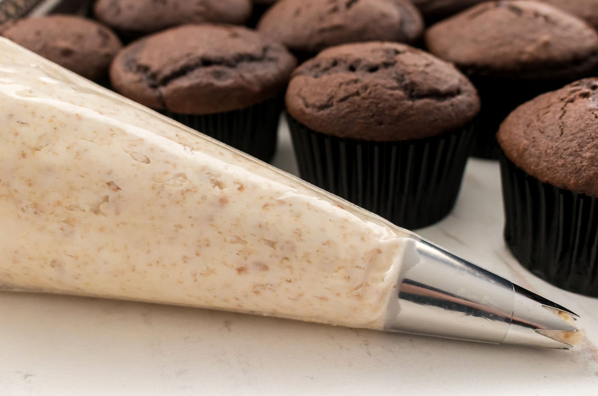 Closeup on a decorating bag filled with Graham Cracker Buttercream Frosting sitting in front of a batch of unfrosted chocolate cupcakes.