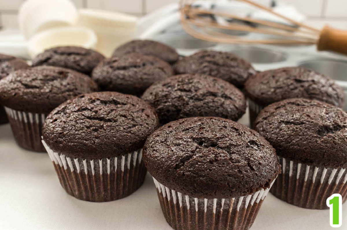 Closeup on a dozen chocolate cupcakes sitting on a white table in front of a cupcake tin and cupcake liners.