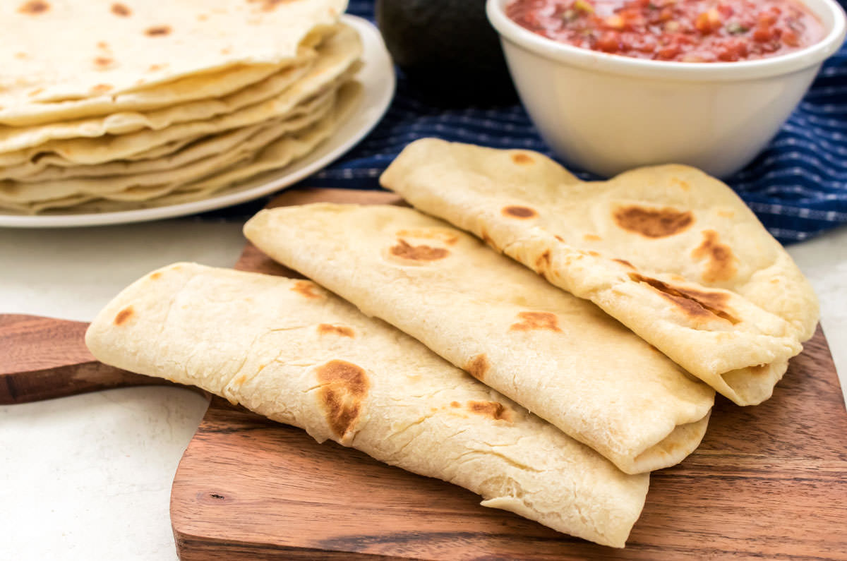 Three Homemade Flour Tortillas, each folded in half, laying on a cutting board in front of a plate of Flour Tortillas and a white bowl filled with salsa.