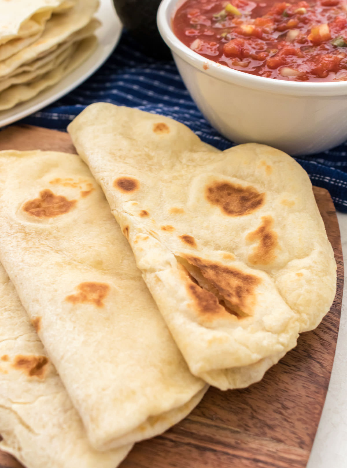 Closeup of three Homemade Flour Tortillas, folded in half, laying on a cutting board in front of a stack of tortillas and a bowl of Pico de Gallo.
