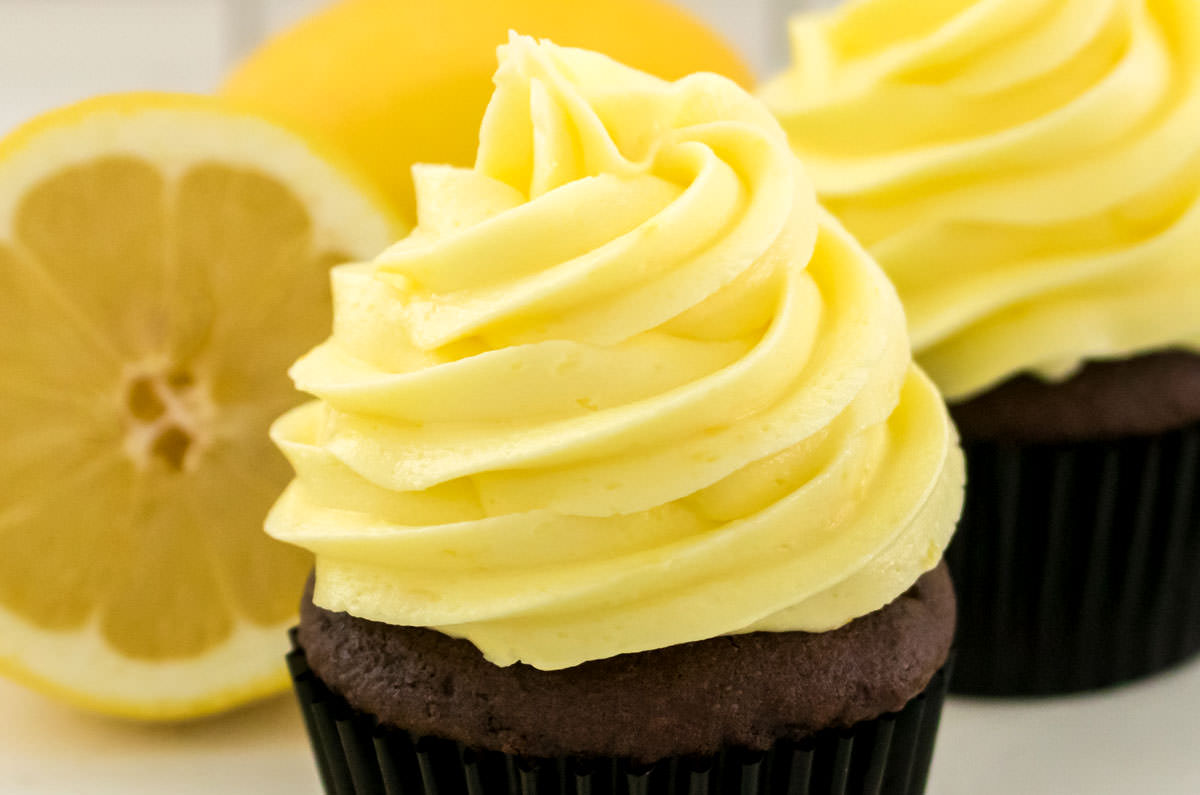 Closeup on two chocolate cupcakes frosted with The Best Lemon Buttercream Frosting sitting next to a fresh lemon.