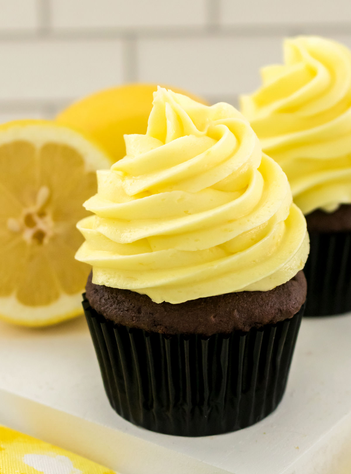 Closeup on a chocolate cupcake topped with The Best Lemon Buttercream Frosting sitting on a white platter next to a fresh lemon.