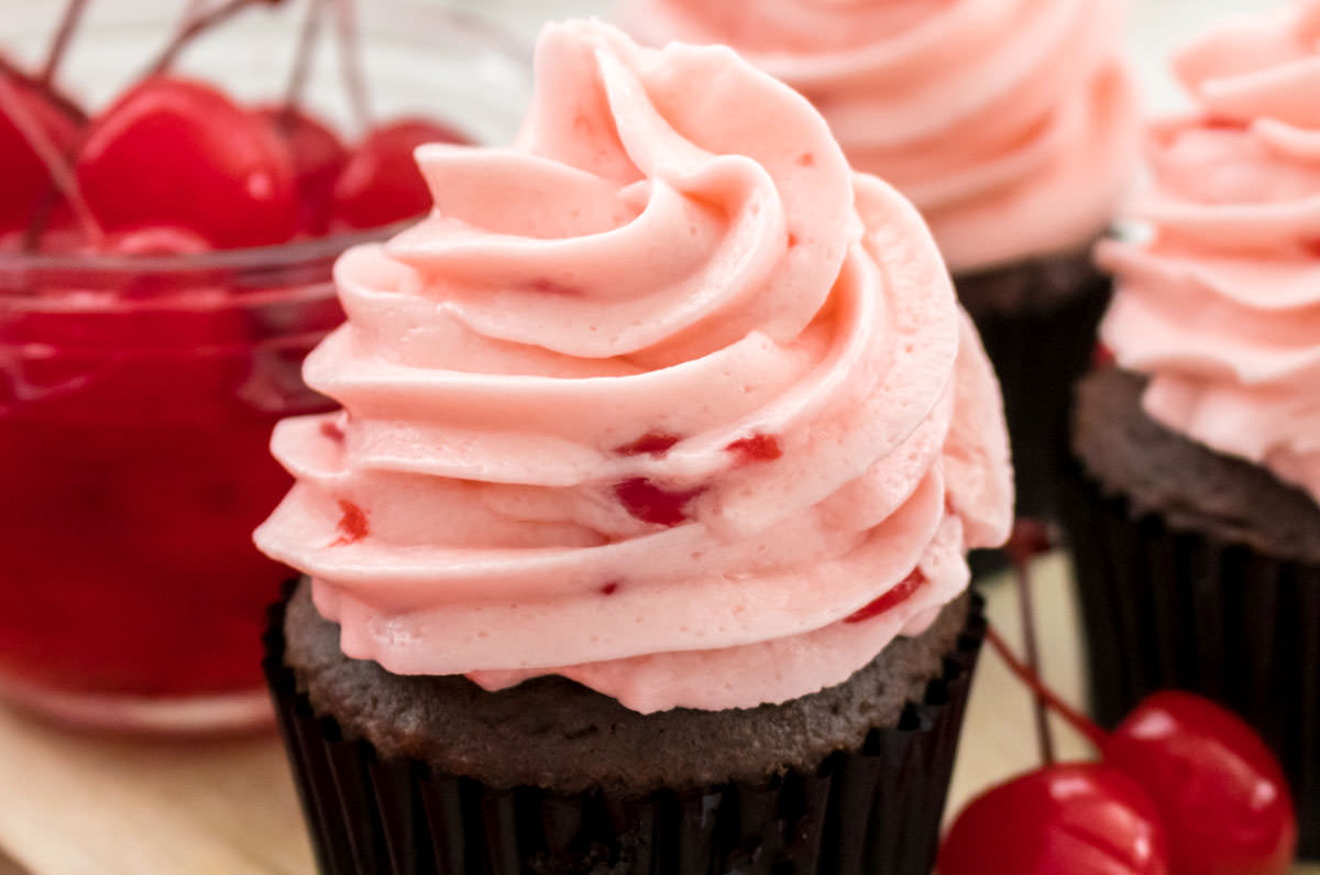 Closeup on three cupcakes topped with The Best Maraschino Cherry Buttercream Frosting sitting next to a glass bowl filled with Maraschino Cherries.