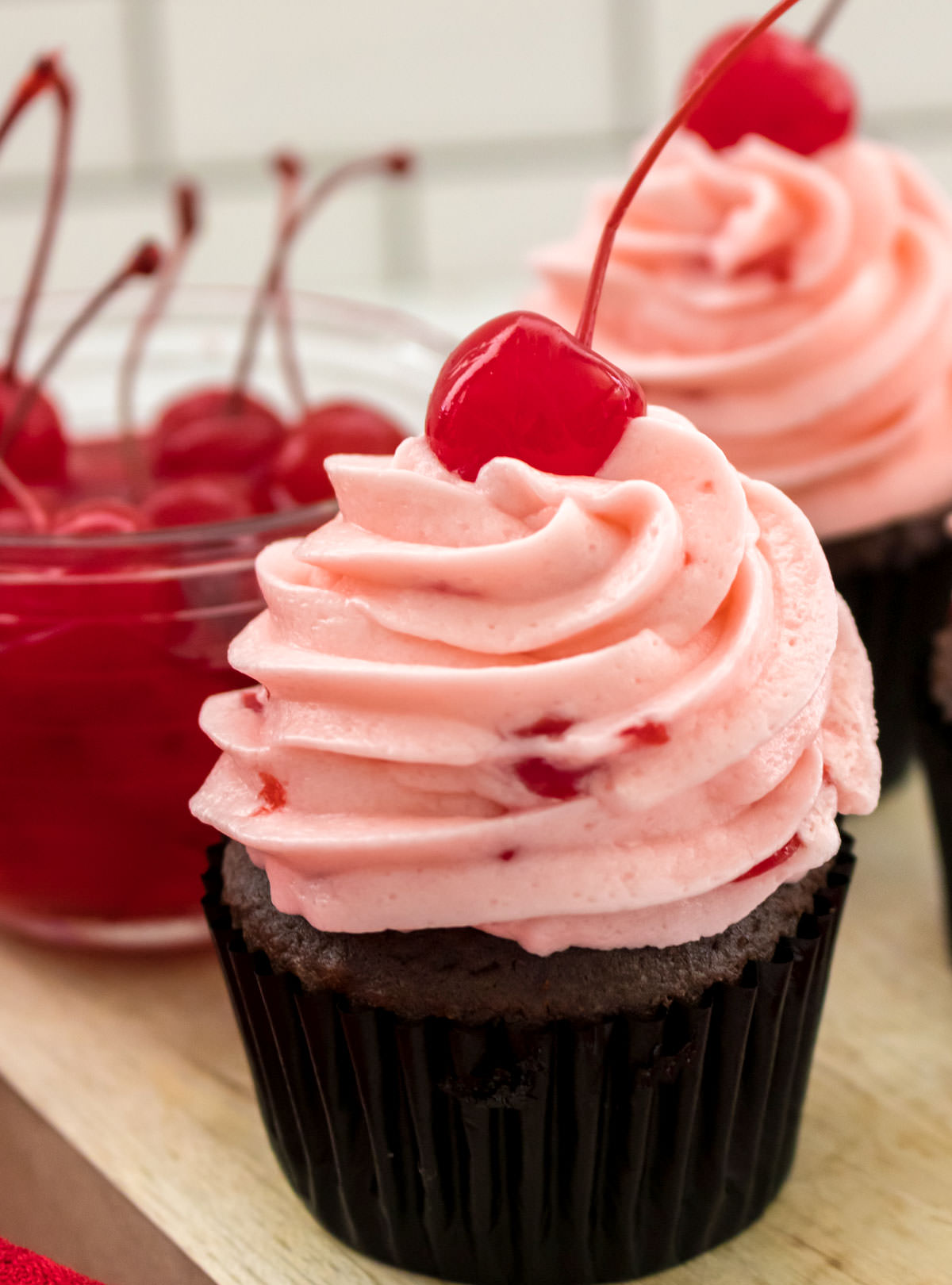 Closeup on a chocolate cupcake topped with The Best Maraschino Cherry Buttercream Frosting sitting next to a glass bowl filled with Maraschino Cherries.