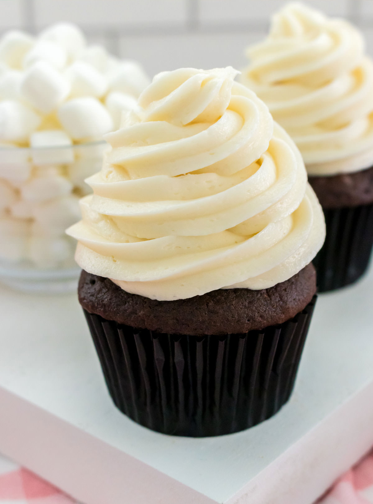 Closeup on two chocolate cupcakes topped with a large swirl of The Best Marshmallow Frosting sitting next to a glass bowl filled with mini marshmallows.