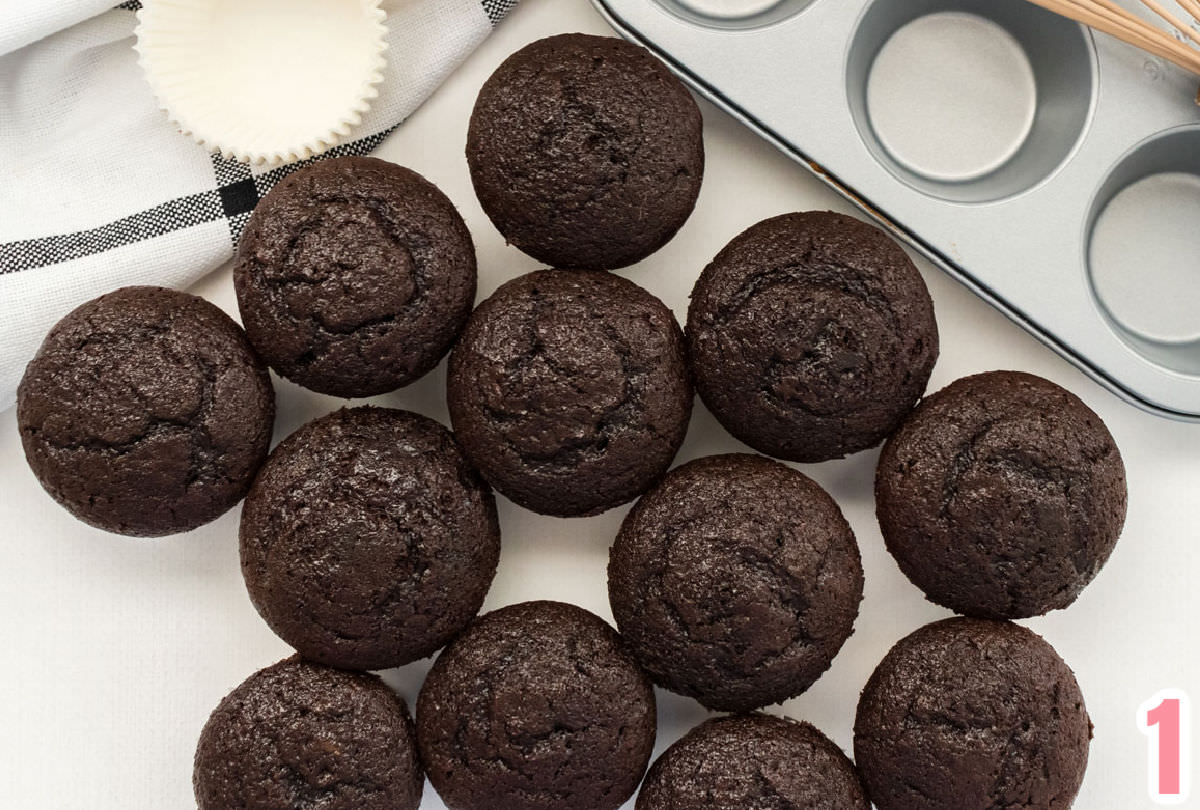 Twelve chocolate cupcakes sitting on a white surface in front of a cupcake pan, cupcake liners and a black and white towel.