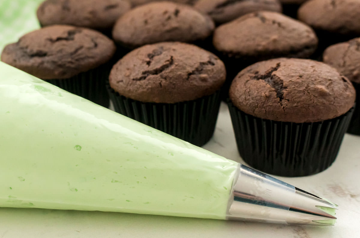 Closeup on a decorating bag filled with Mint Frosting sitting in front of a batch of unfrosted chocolate cupcakes.